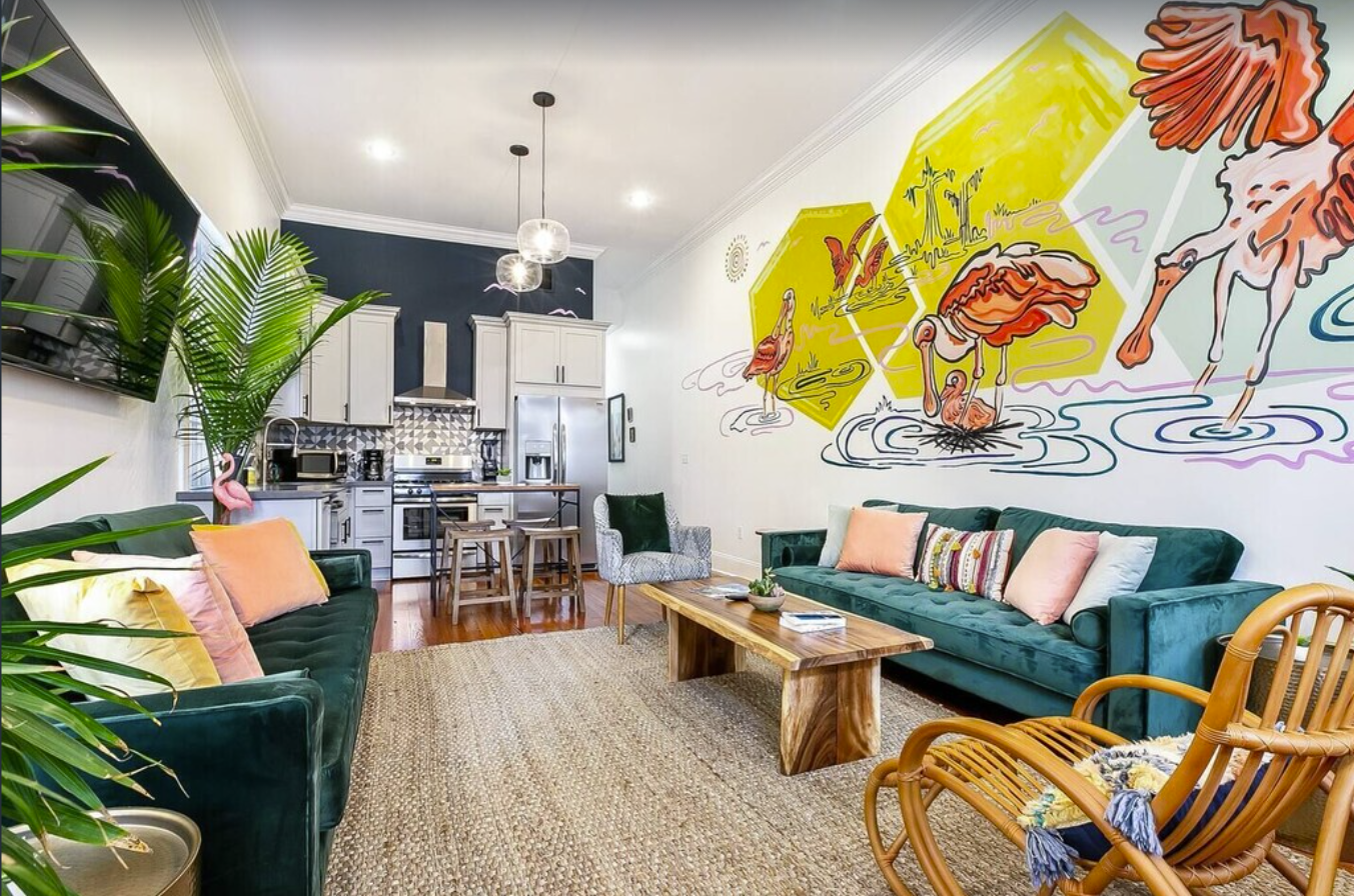 An interior image of a living room in a New Orleans townhome. The white walls are painted with colorful artwork. Two emerald green couches sit across from each other, surrounded by other furniture, house plants, and a wall-mounted TV