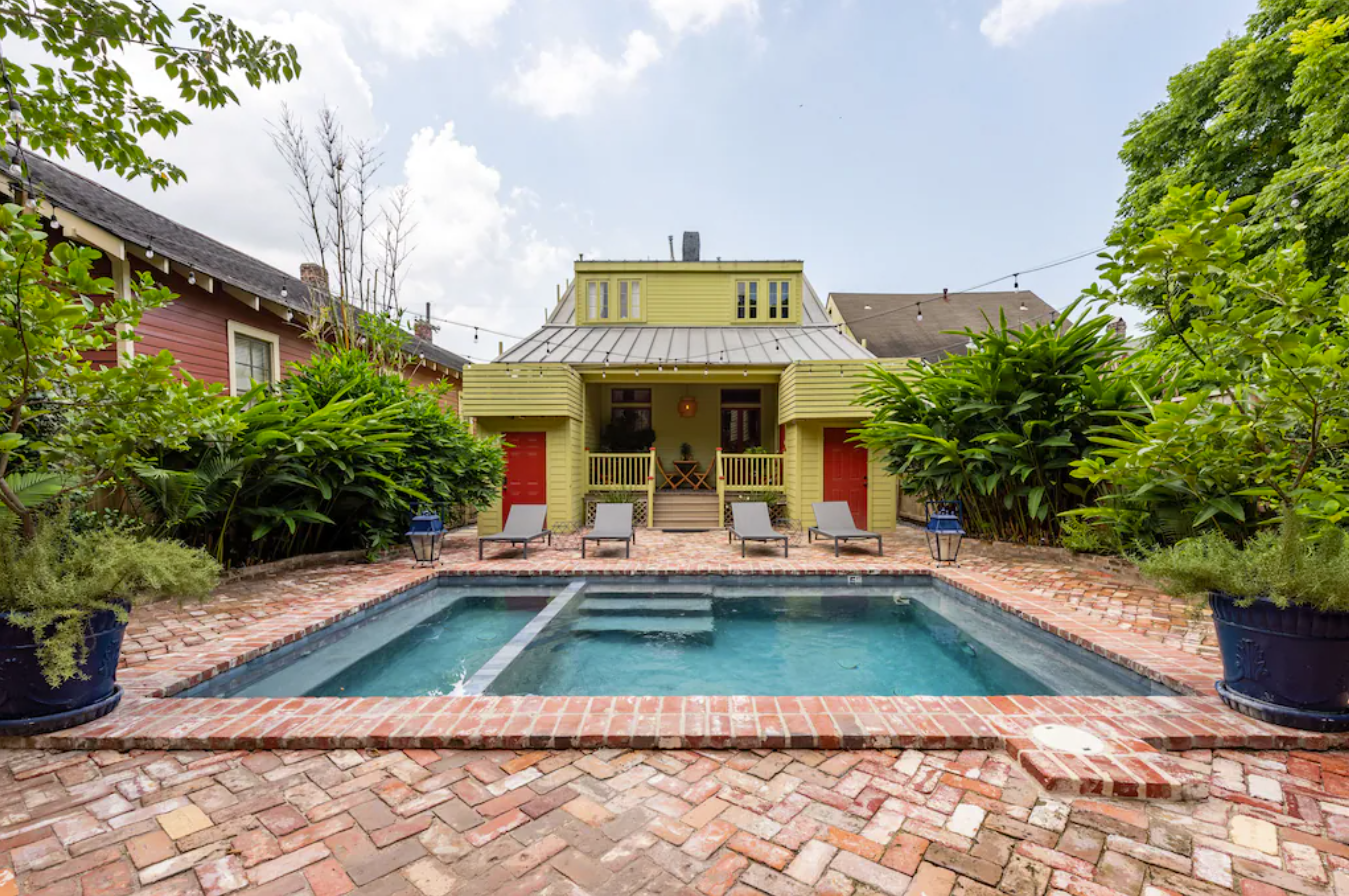 An exterior shot of a backyard with a brick-paved patio around an in-ground pool. In the background is a yellow house. The backyard is surrounded by walls of greenery, providing plenty of privacy.