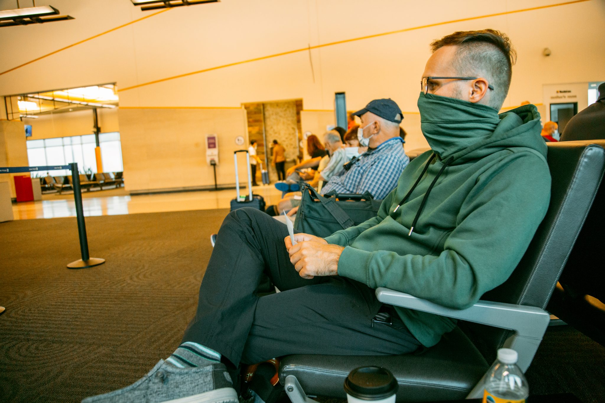 A man sits in airport chairs, wearing a green hooded sweatshirt with a turtleneck mask pulled over his face.