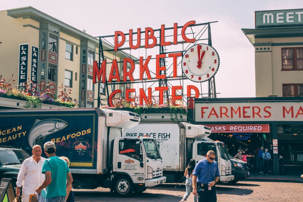 Pike Place Market sign and clock with fish trucks parked in front on a sunny day