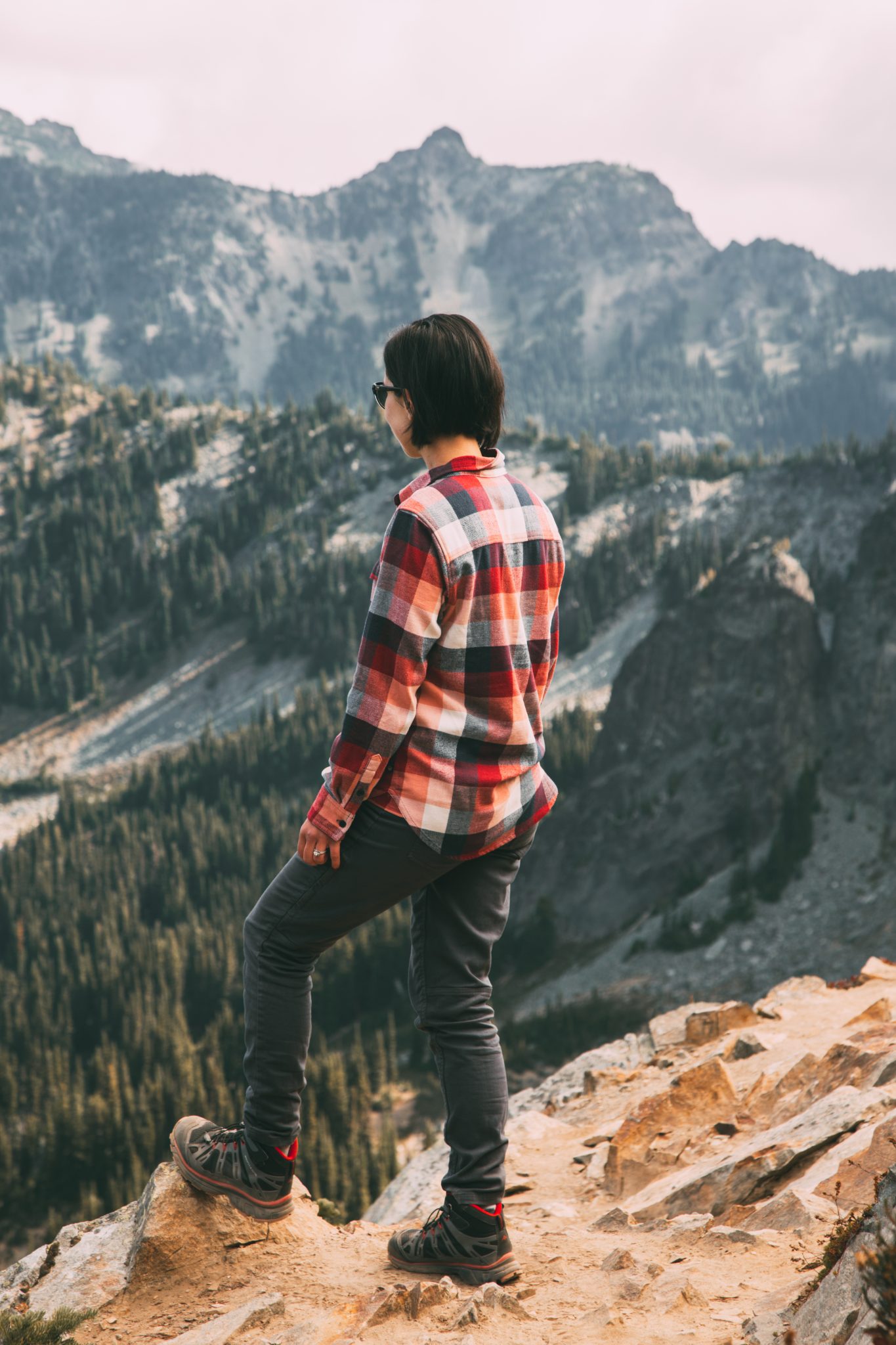 A woman poses on a mountain ridge, overlooking the scenery. She's wearing a red, white, and black plaid shirt and black hiking pants.