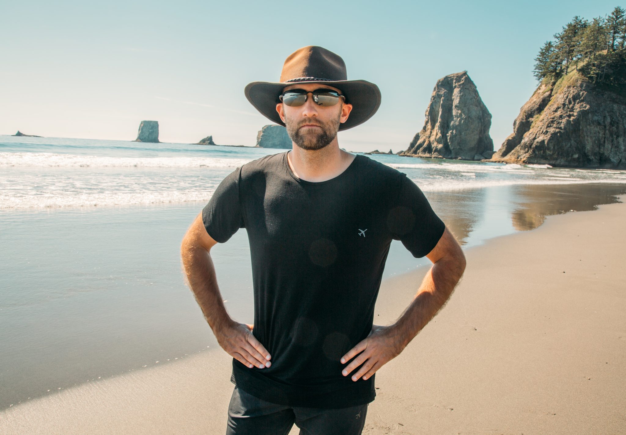 A man poses on a beachfront with his hands on his hips, looking at the camera. He's wearing a hat, sunglasses, and a black t-shirt.