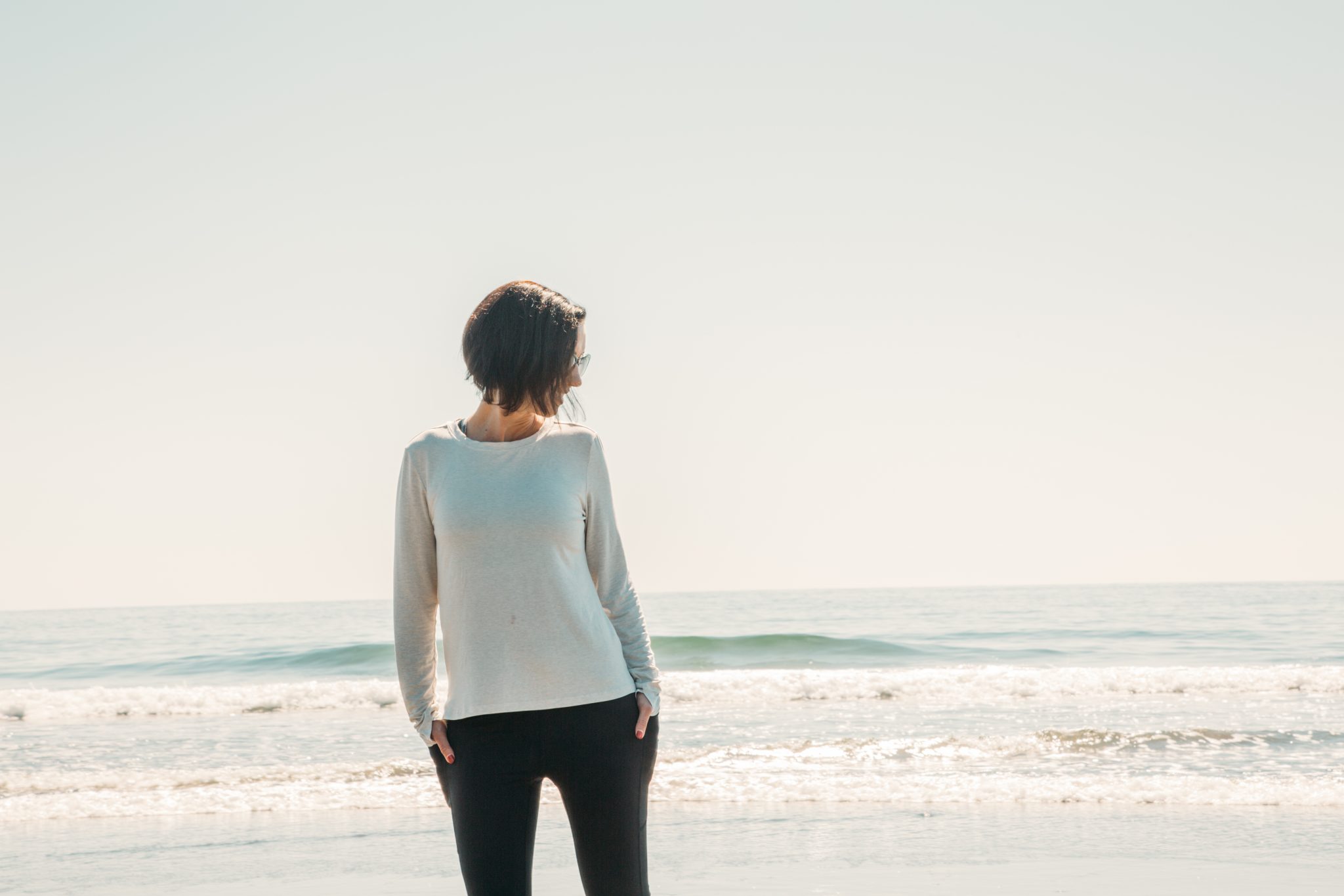 A woman poses on a beach in front of the ocean with waves cresting behind her. She's wearing a long-sleeved white shirt and black leggings from sustainable travel clothing brands.