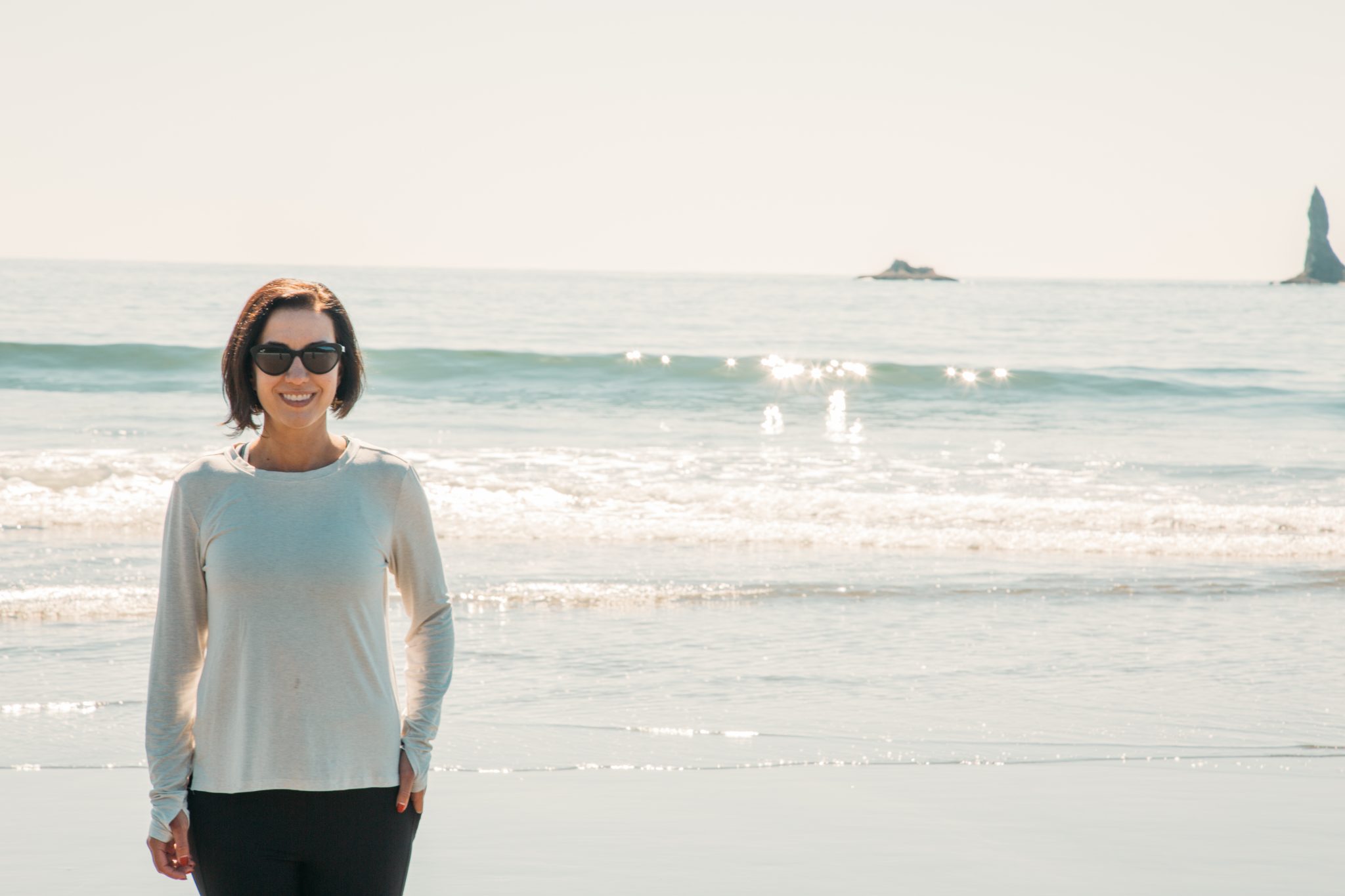 A woman stands in front of the ocean, waves crashing behind her, smiling at the camera. She's wearing a white long sleeve shirt and black pants.