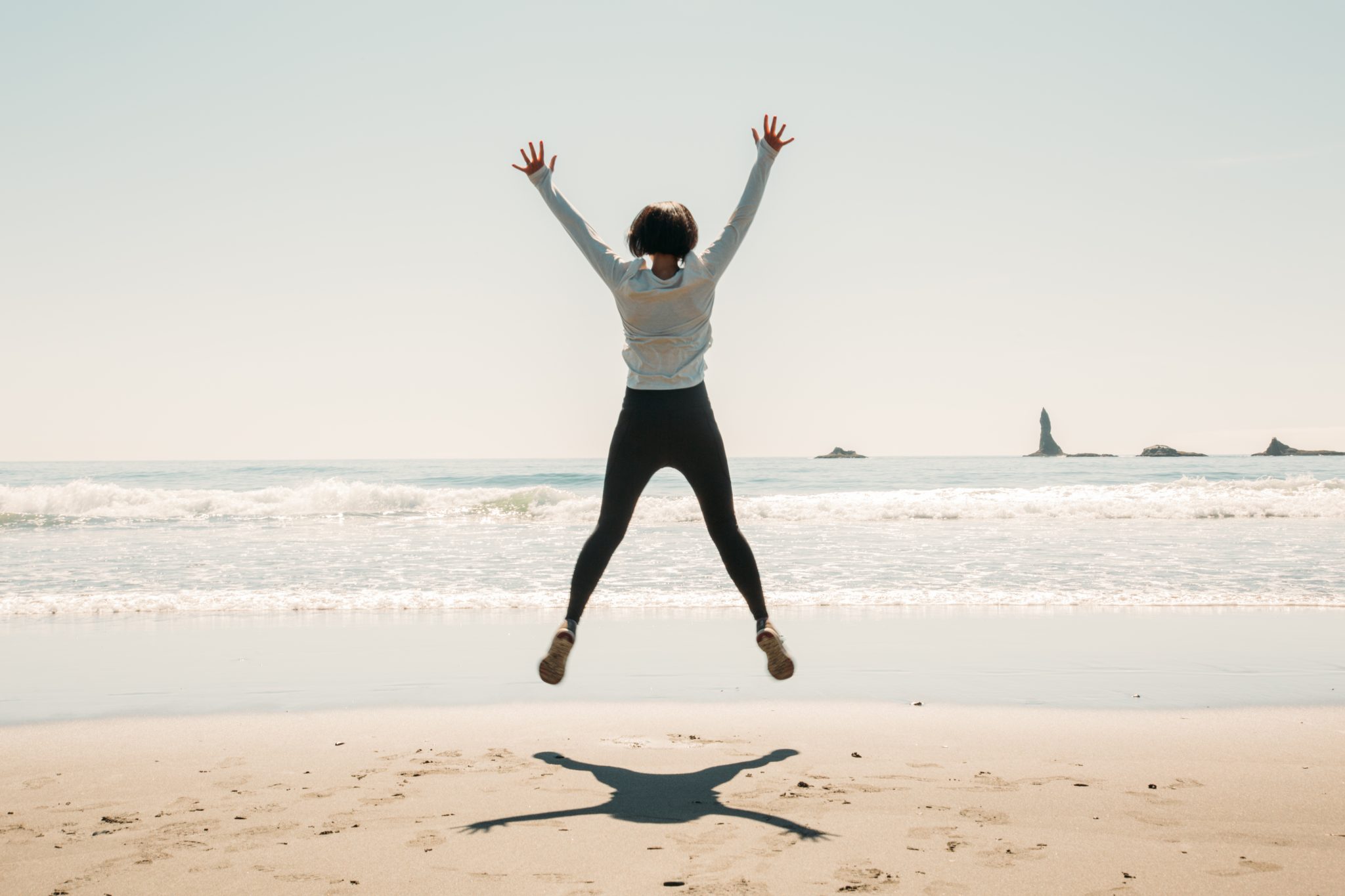 A woman jumps in the air, arms spread wide, with her back to the camera, on the shoreline in front of the ocean on a sunny, clear day.