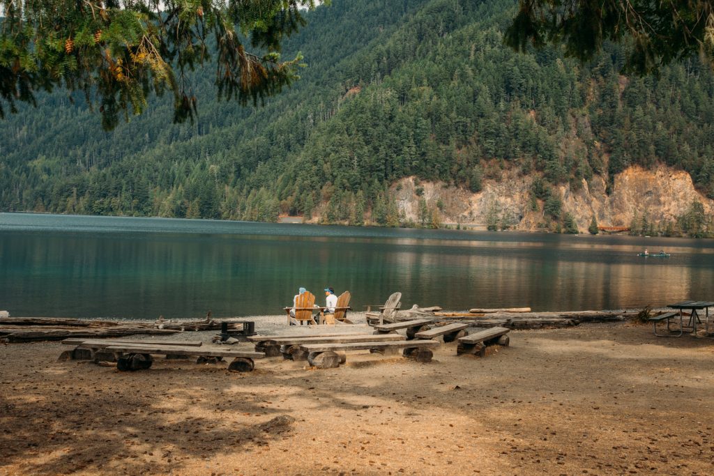 A couple sits in wooden chairs looking at a beautiful lake and mountain with pine trees at Lake Crescent Lodge