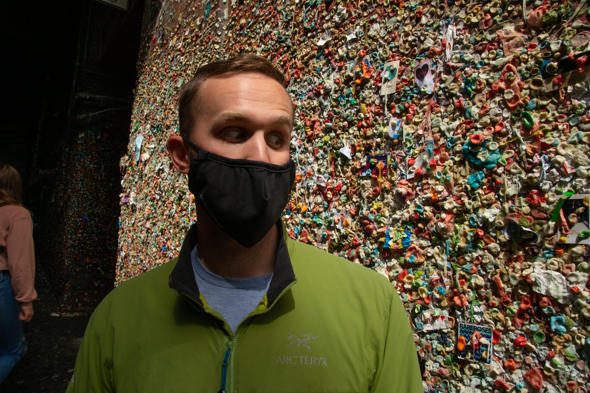 A man stands in front of the infamous gum wall in Seattle, wearing a face mask and green pull-over jacket.