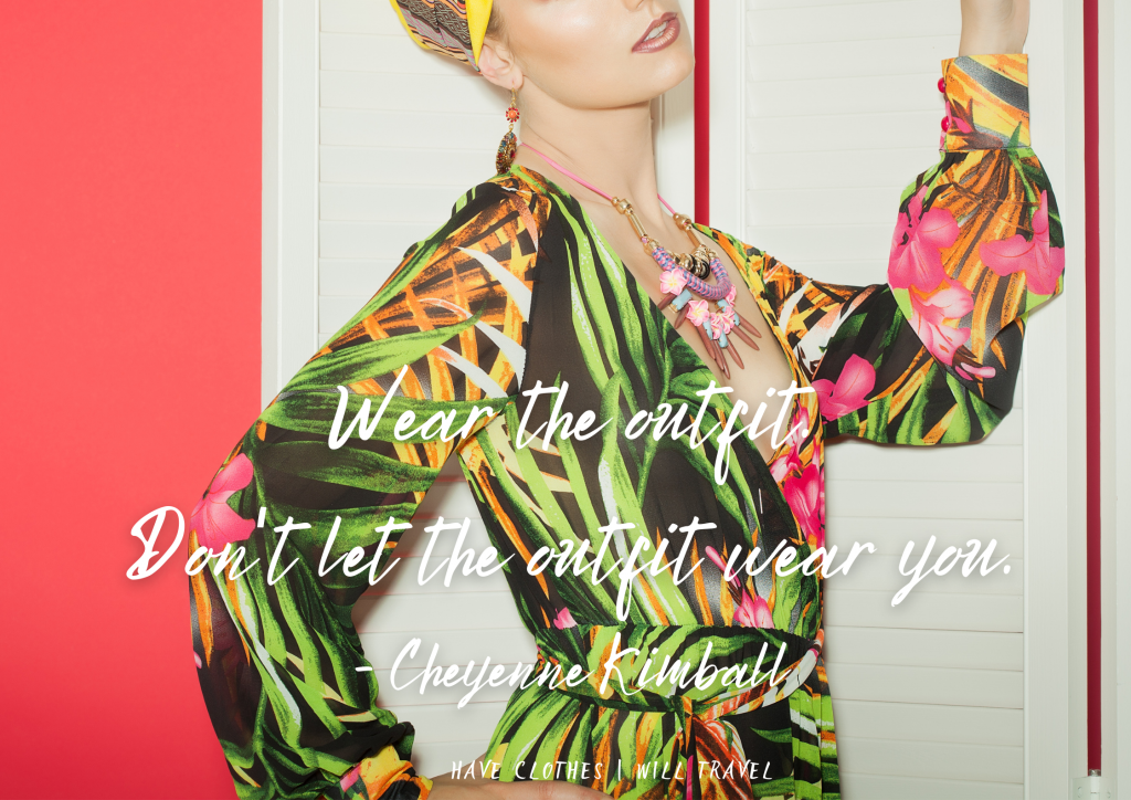 A model stands in front of a white door and red wall. She's wearing a tropical patterned dress and colorful accessories. White text overlayed on the image says "Wear the outsit, don't let the outfit wear you. - Sheyenne Kimball