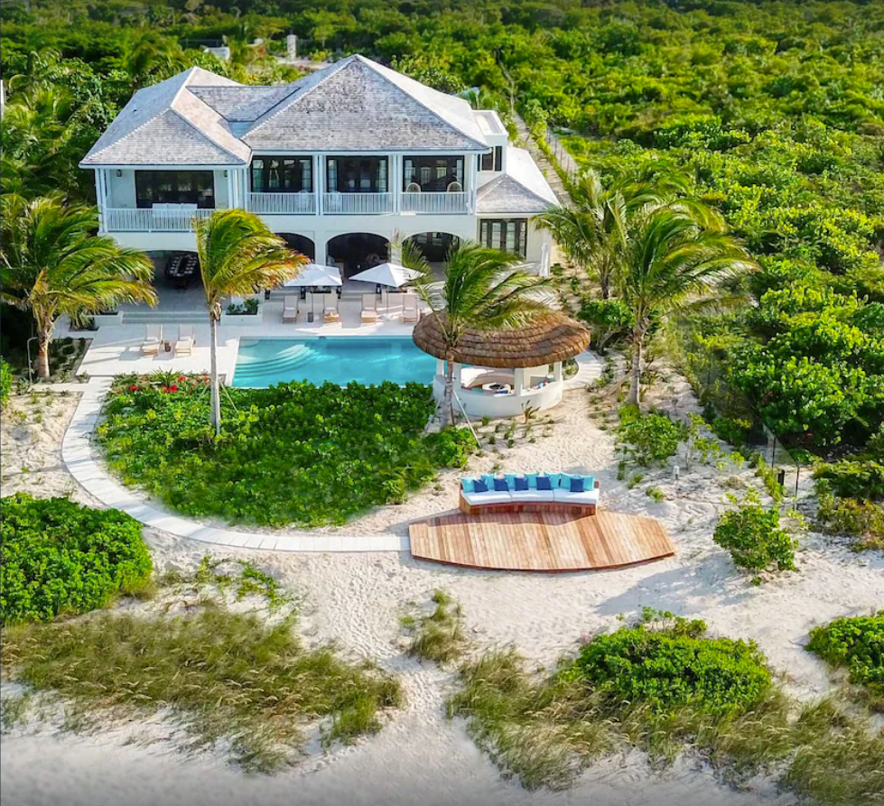 An aerial view of a large, secluded tropical villa in Turks and Caicos features a two-story home with wrap-around porches, a private pool, multiple lounging areas, and direct access to a sandy beach.