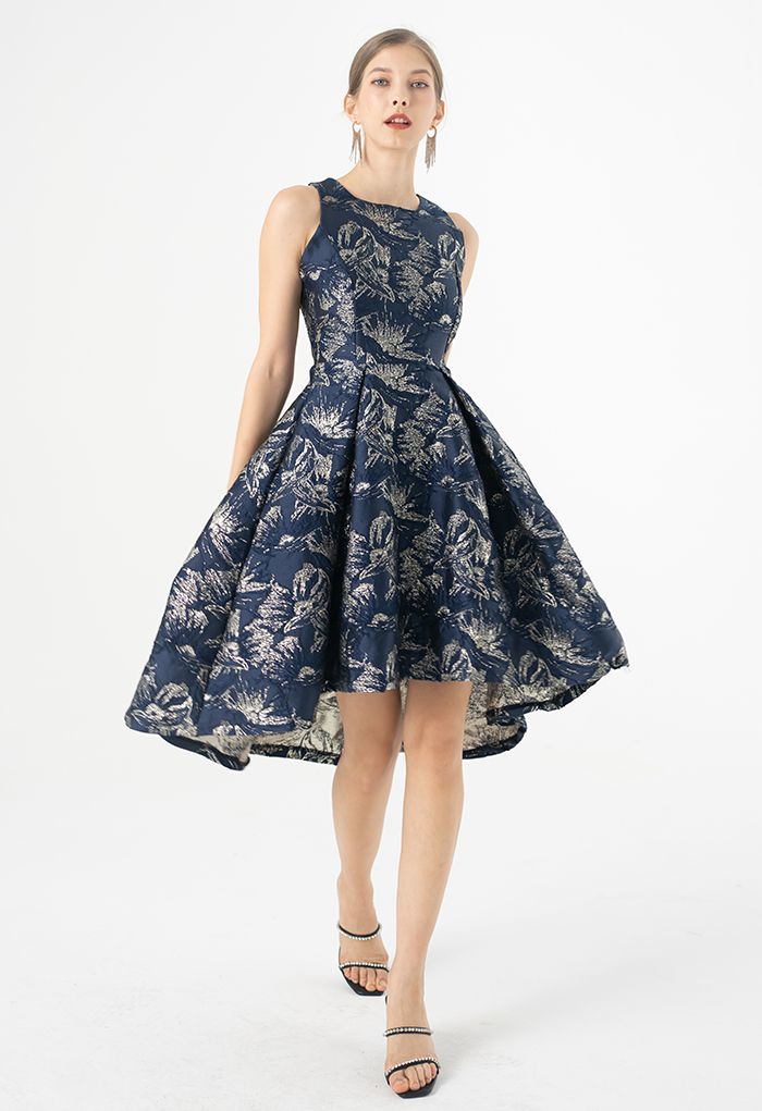A white female model wears a navy cocktail dress with a silver shimmer magnolia pattern. The knee-length a-line skirt is pleated from the waist down, and the model is wearing strappy black heels.