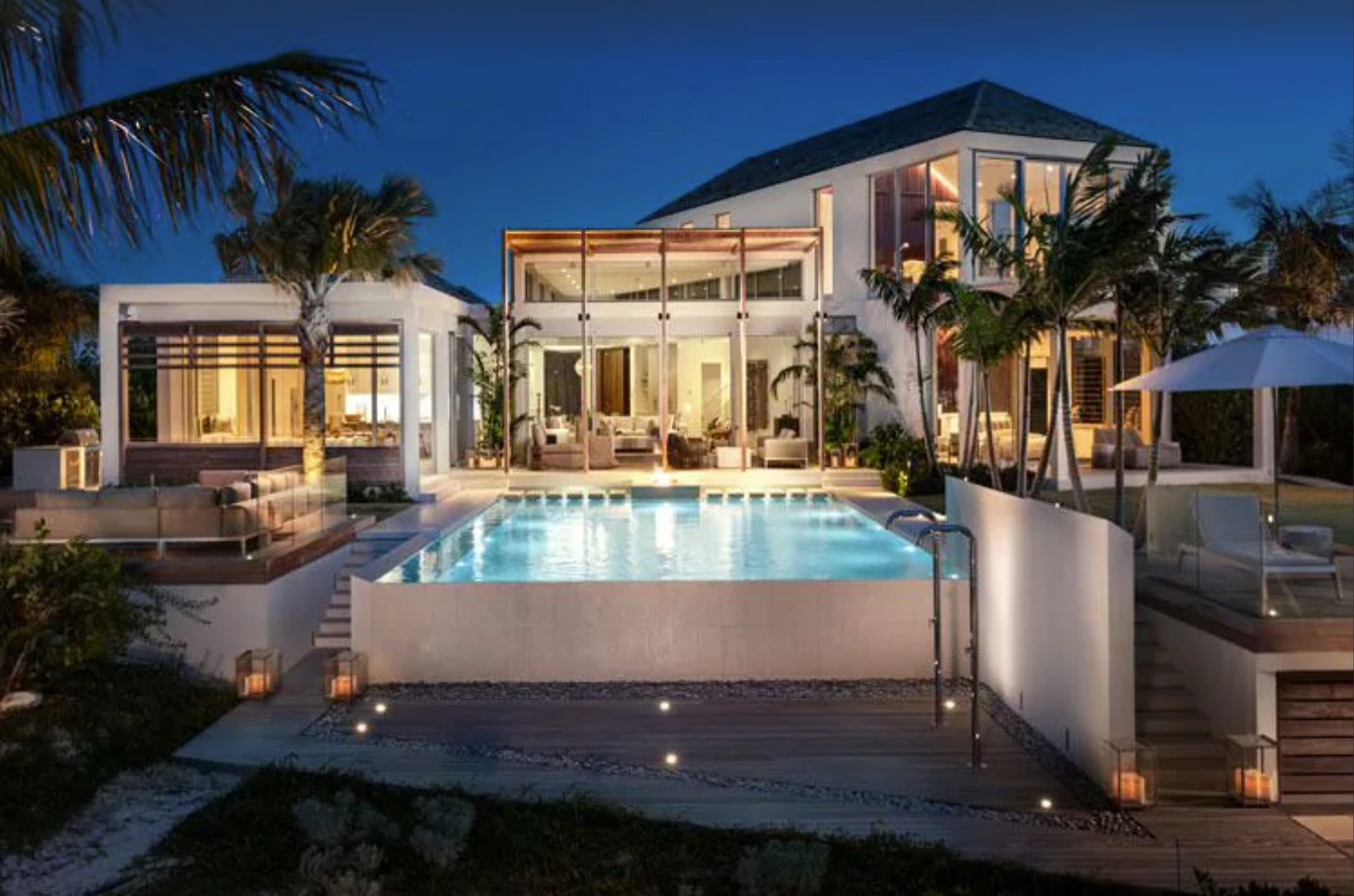 An exterior image of a Turks and Caicos rental villa at night. The private pool and outdoor areas are lit with soft pathway lighting, and the two-story home glows from within, with floor-to-ceiling 360-degree windows.