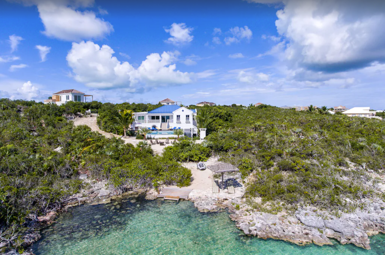An aerial view of a private home in Turks and Caicos. The home is white with a blue roof and is secluded, surrounded by trees. There's oceanfront access and a private beach and the edge of the porperty.