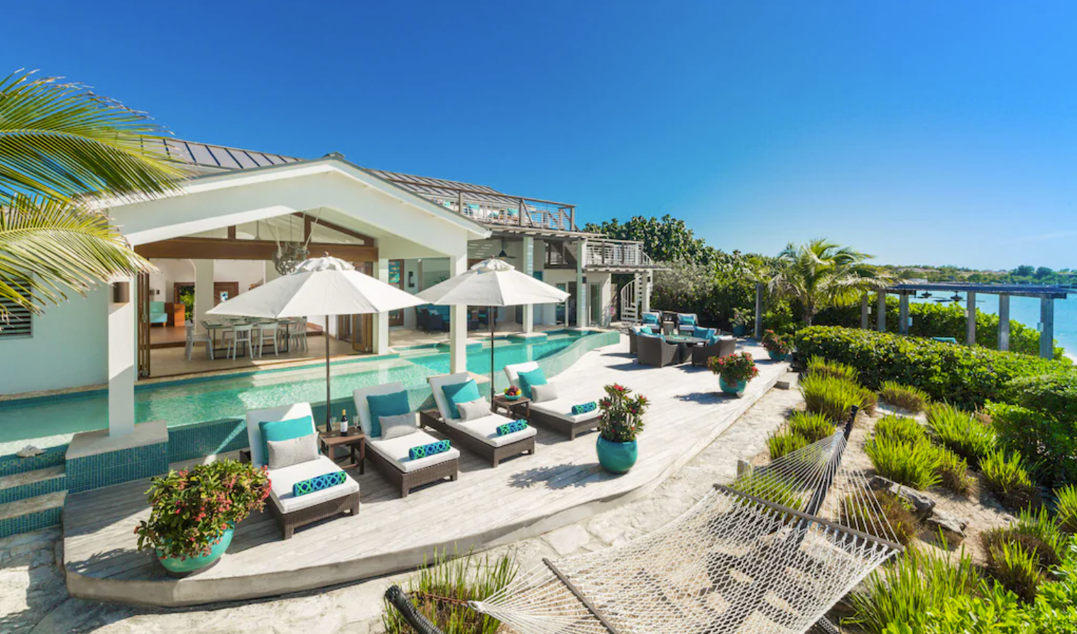 An aerial view of a luxury Turks and Caicos vacation rental with a private pool. Shaded lounge chairs overlook the ocean from a private patio with a gorgeous pool, and a two-story home with floor-to-ceiling glass panel windows.