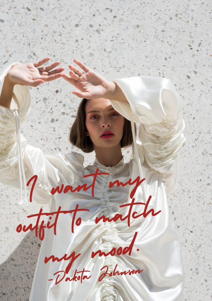 A model is wearing a ruched white satin dress and stands in front of a stone wall with her hands raised above her red. Red text over the image says, "I want my outfit to match my mood. - Dakota Johnson"
