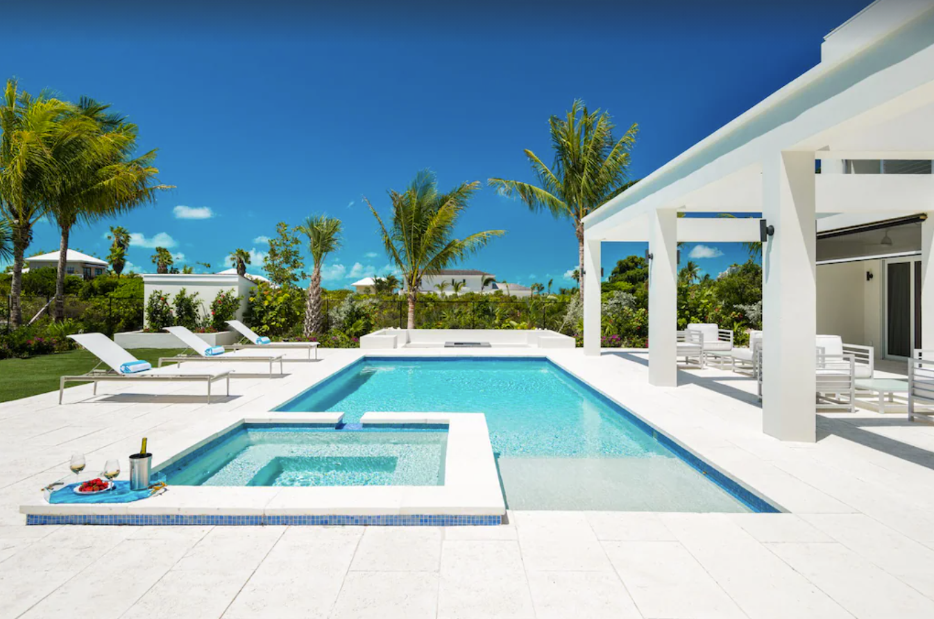 The outdoor private pool area of a Turks and Caicos rental villa features a pool and jacuzzi, lounge chairs, covered patio areas surrounded by privacy landscaping.