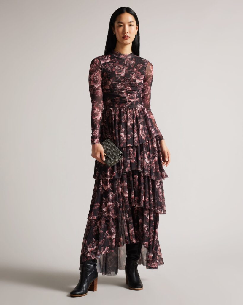 An Asian female model wears a long sleeve dress with a light all over floral print. The skirt flows in tiered layers, and the dress has a mock turtleneck neckline. 