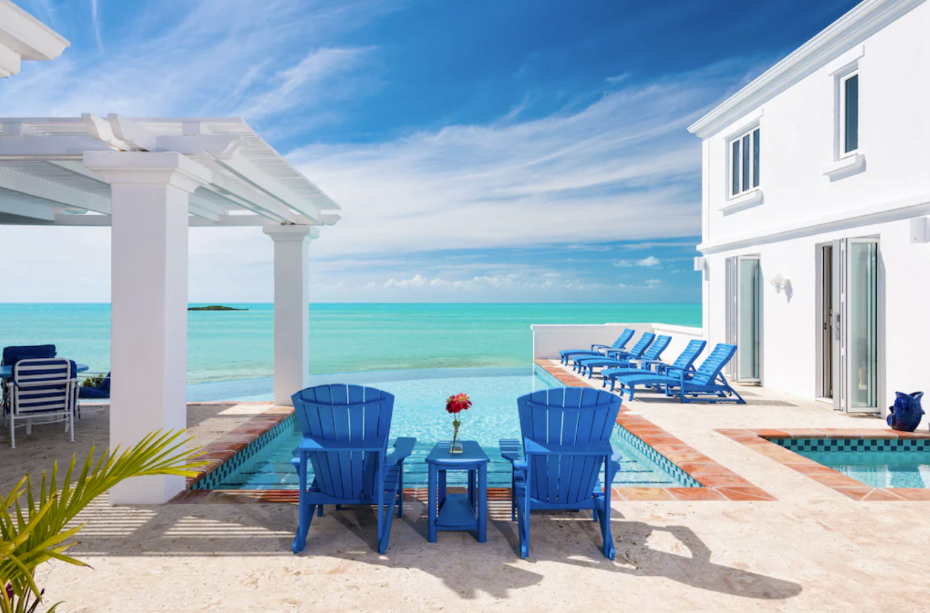Two blue Adirondack chairs sit at the edge of a private pool overlooking the ocean. To the left is a covered patio area and to the right, French doors that open up into villa bedrooms.