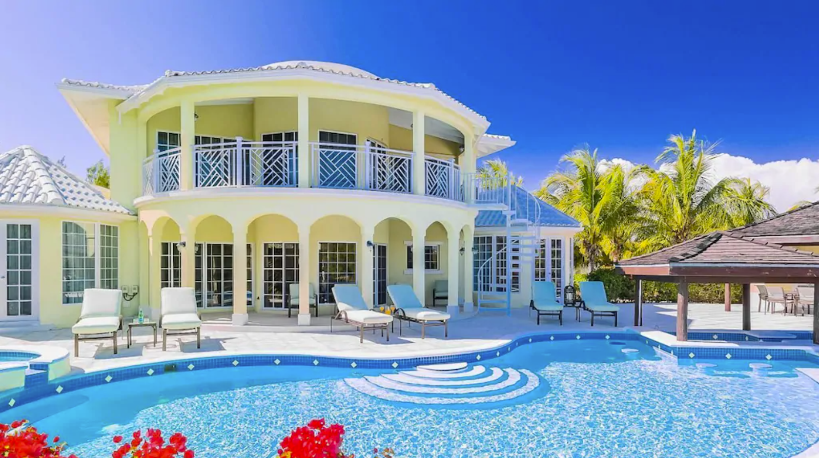 An exterior image of a stunning two-story Turks and Caicos villa. The exterior of the home is a quaint yellow with white roofing and trim, a second story balcony, and covered porch. The private patio has a large pool and shaded and unshaded sunning areas.