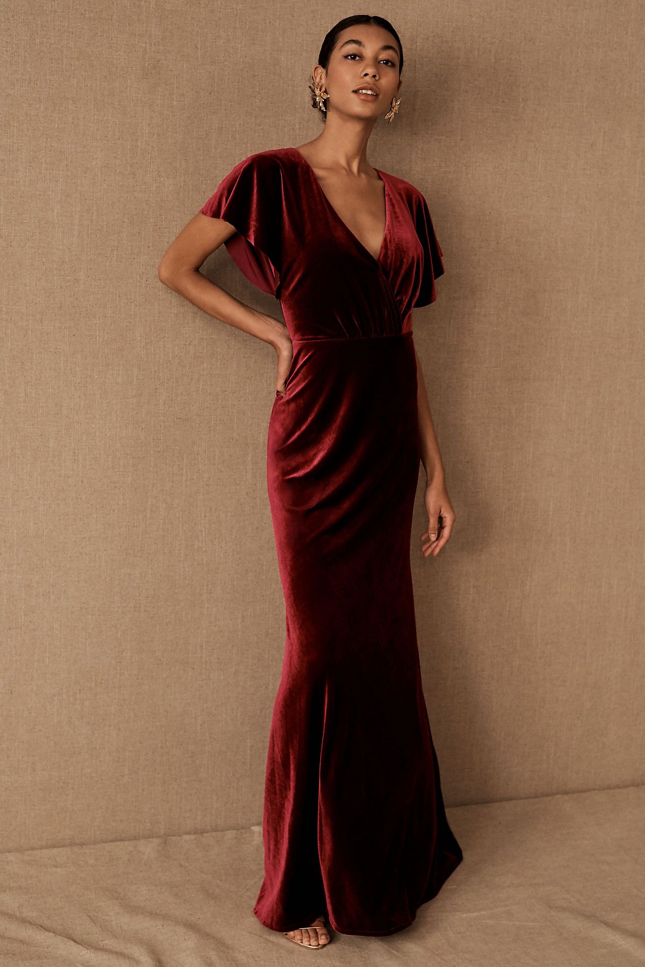 A young darker-skinned model poses against a tan background, wearing floor-length deep red velvet dress with a deep v neckline and flutter sleeves.