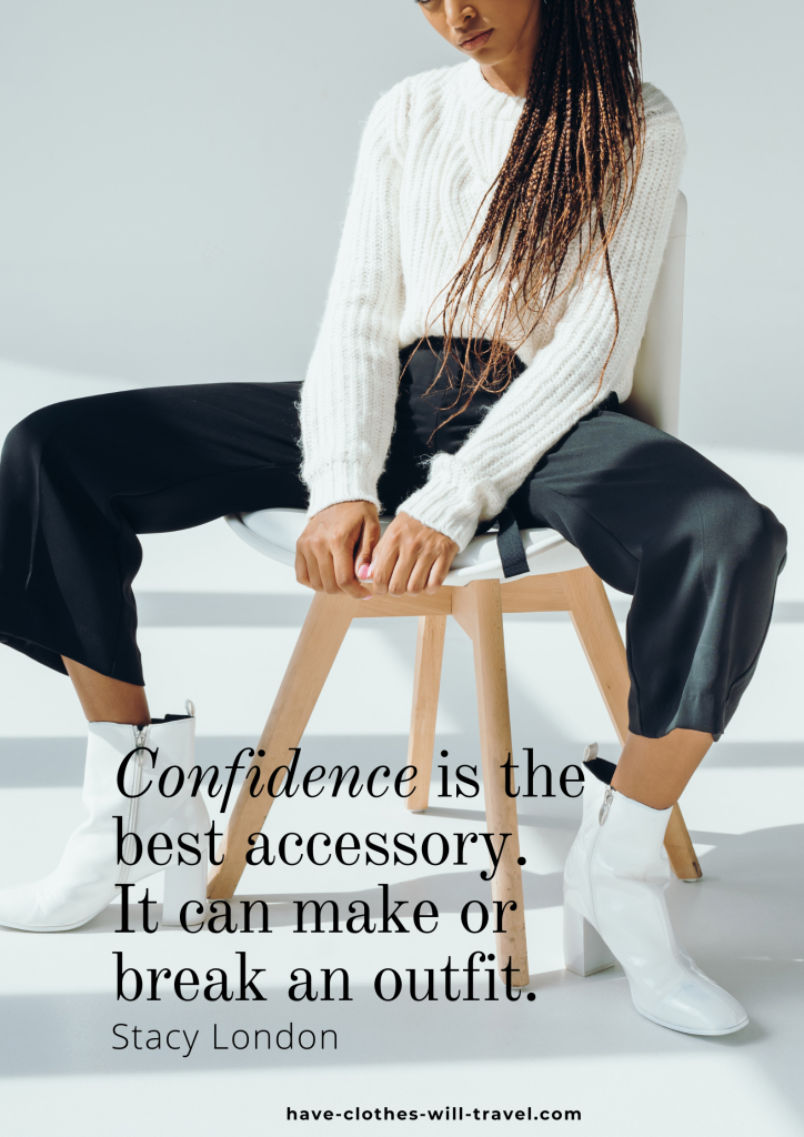 A young African American woman with long, braided hair sits on a white wooden chair in a white room. She's wearing black slacks and a white cable knit sweater and white heeled boots. Black text overlayed on the image reads "Confidence is the best accessory. It can make or break an outfit. - Stacy London" 