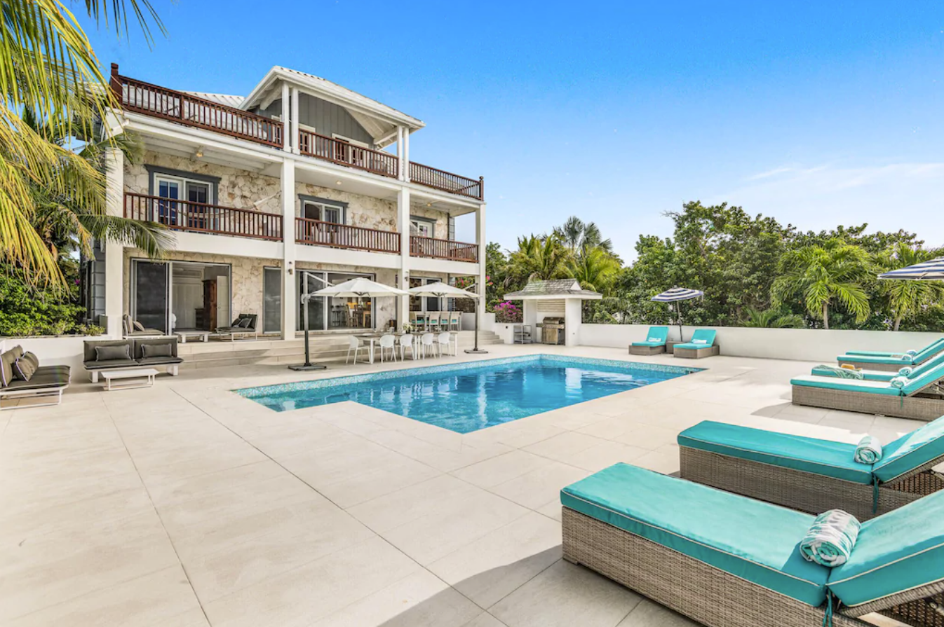 An exterior image of the private pool and patio space of a villa in Turks and Caicos. The two-story home has wrap-around balconies that overlook a private pool surrounded by lounge chairs.