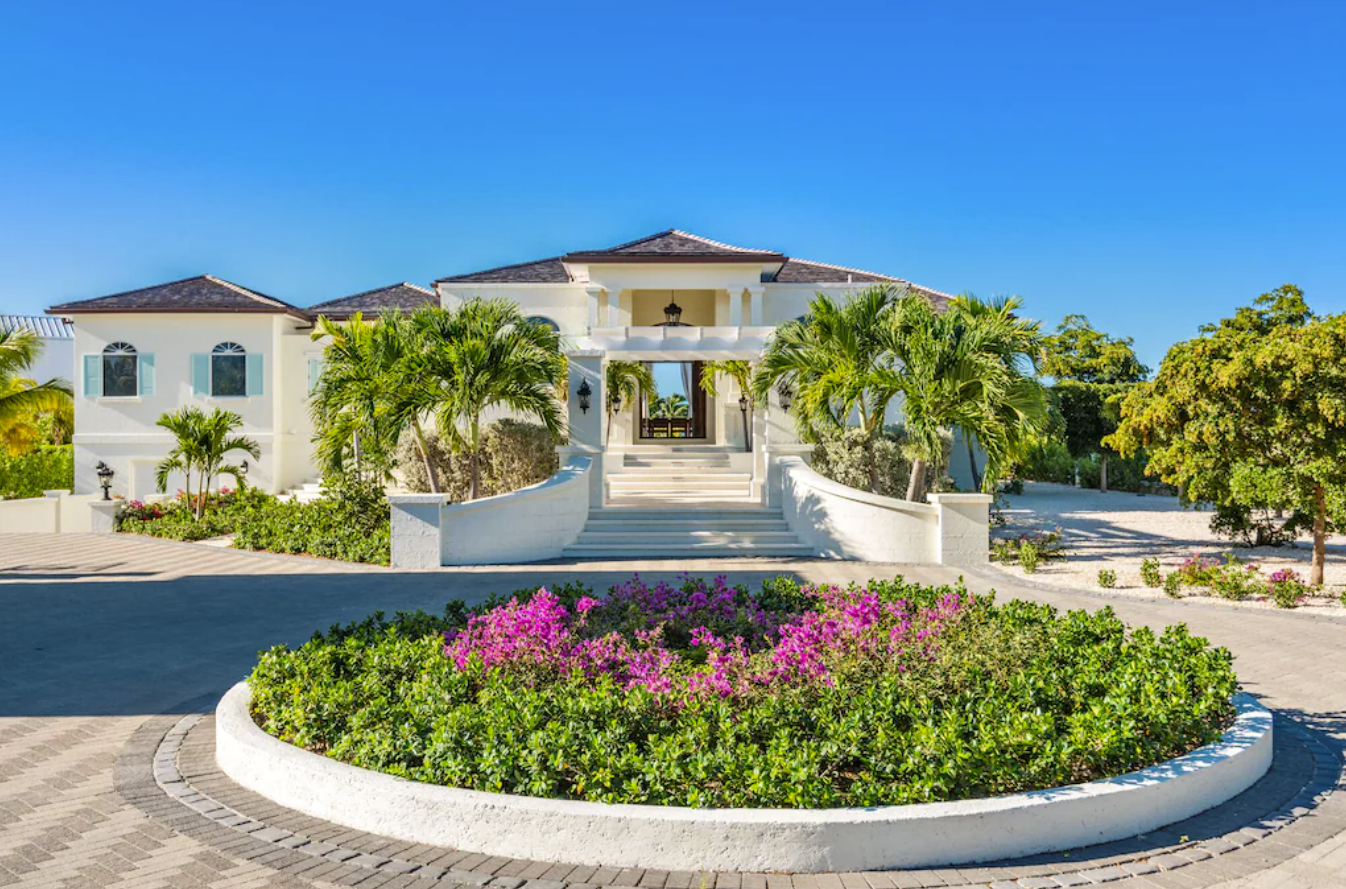An exterior image of the grand front entrance to a luxury Turks and Caicos rental villa. A long round-about driveway leads up to a grand staircase, landscaped with palm trees and tropical plants. 