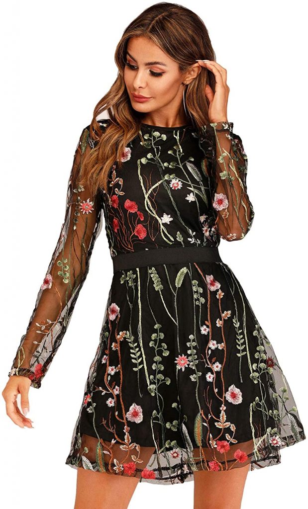 A white female model wears a cocktail length black dress with an all-over colorful embroidered floral pattern. The embroidery continues down the sheer full length sleeves.