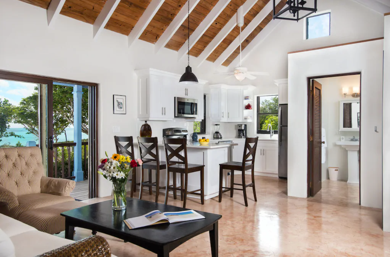 The inside of a quaint Turks and Caicos rental villa features a small kitchen and breakfast bar, tall, open-air ceilings, a small living room areas, and clear ocean view.