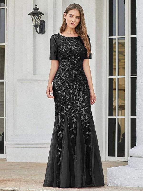 A white female model wears a floor-length black gown with a black sequined leaf pattern. The mermaid-style gown has short sleeves and a flared tulle bottom.