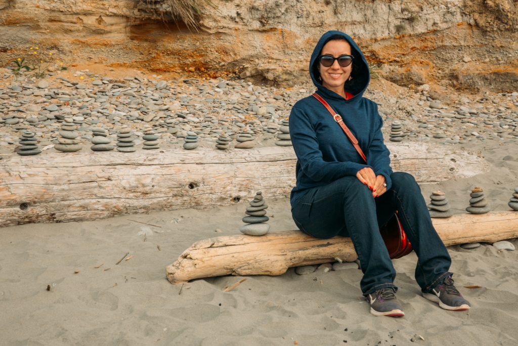 A woman sits on a log surrounded by stacked stone piles. She's wearing a navy blue Aviator Merino wool hoodie from a sustainable travel clothing brand.