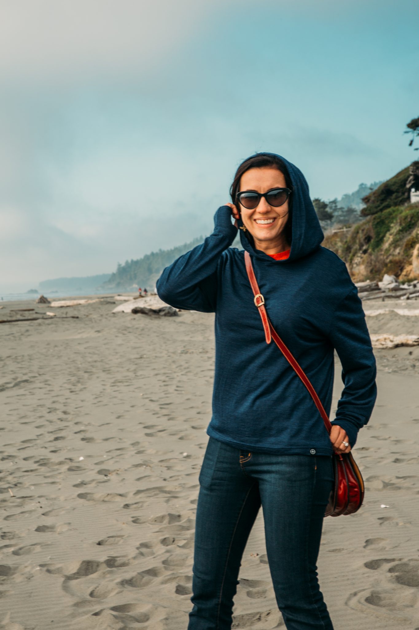 A woman poses on the beach on a cold, cloudy day, wearing a navy blue hooded sweatshirt and jeans.