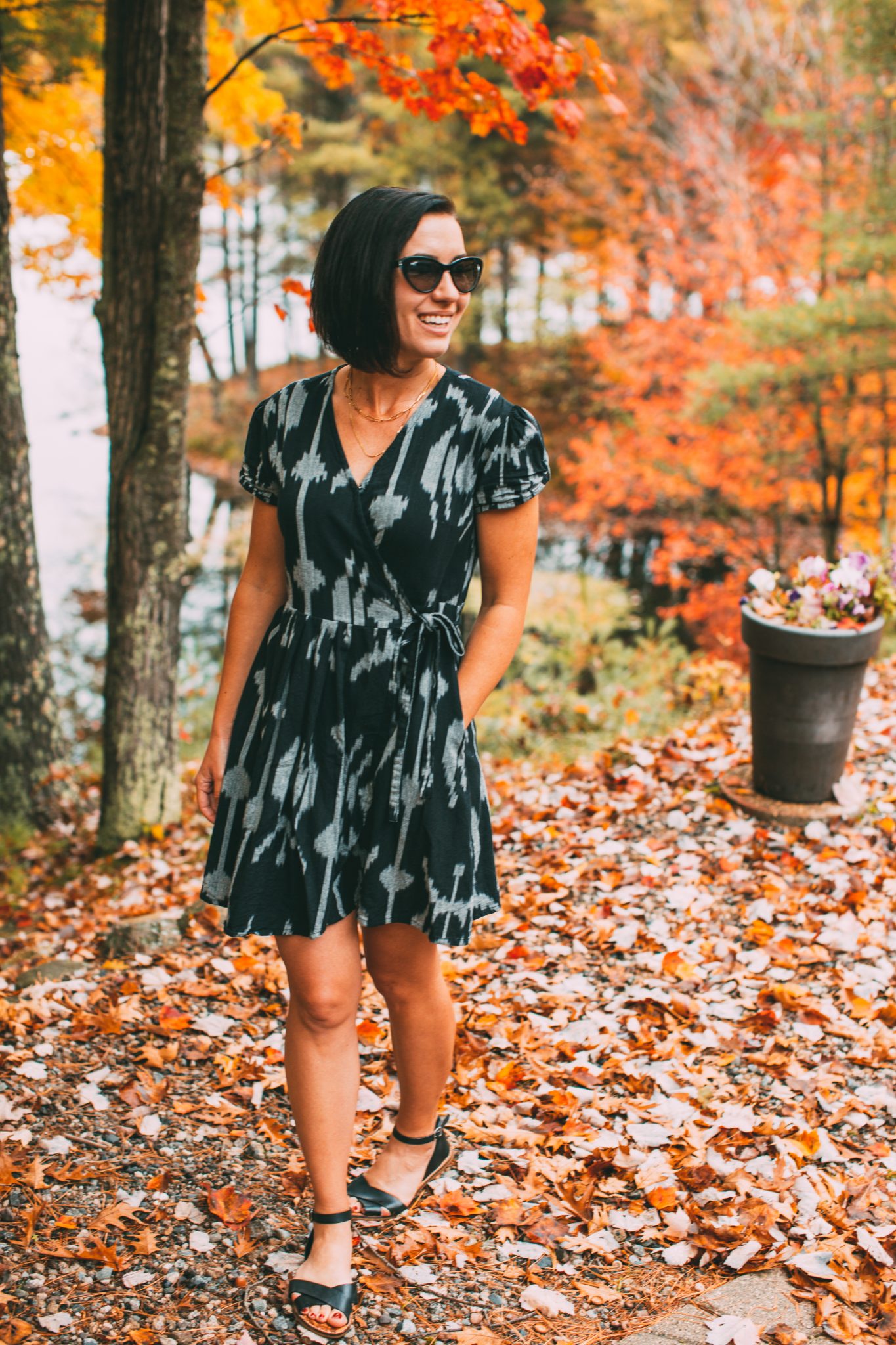 A young woman with short brown hair poses in a colorful Fall landscape, with fallen leaves on the ground, and orange and yellow trees in the background. She's wearing a black and white patterned dress, sustainably made by the fashion brand Passion Lillie.