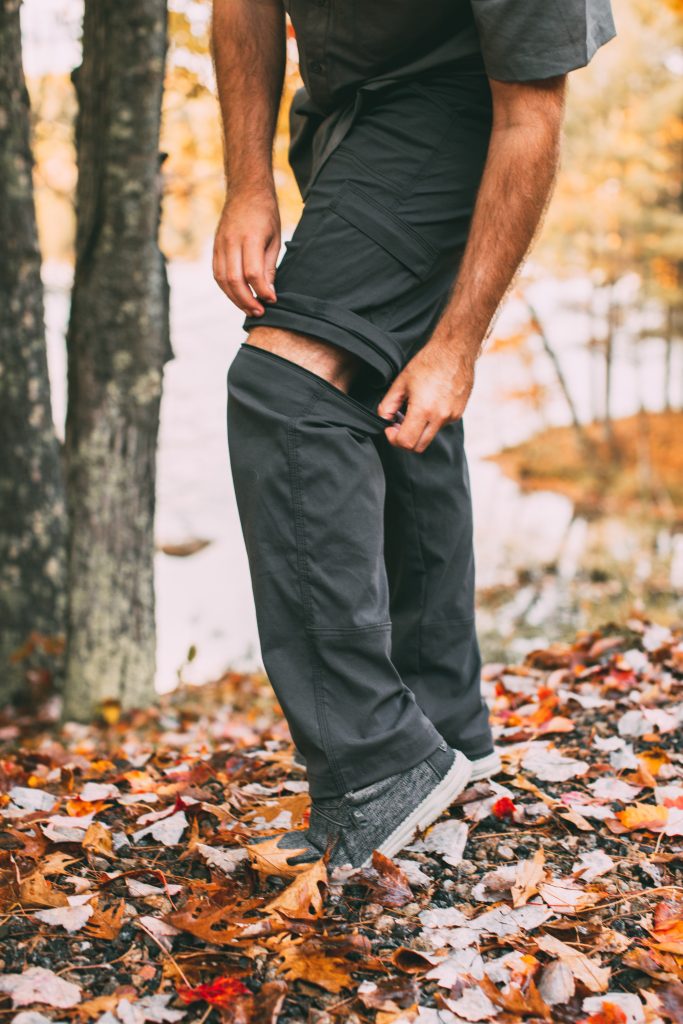 A man poses on leaf-covered ground, demonstrating how his travel pants convert from pants to shorts by unzipping at the knee.