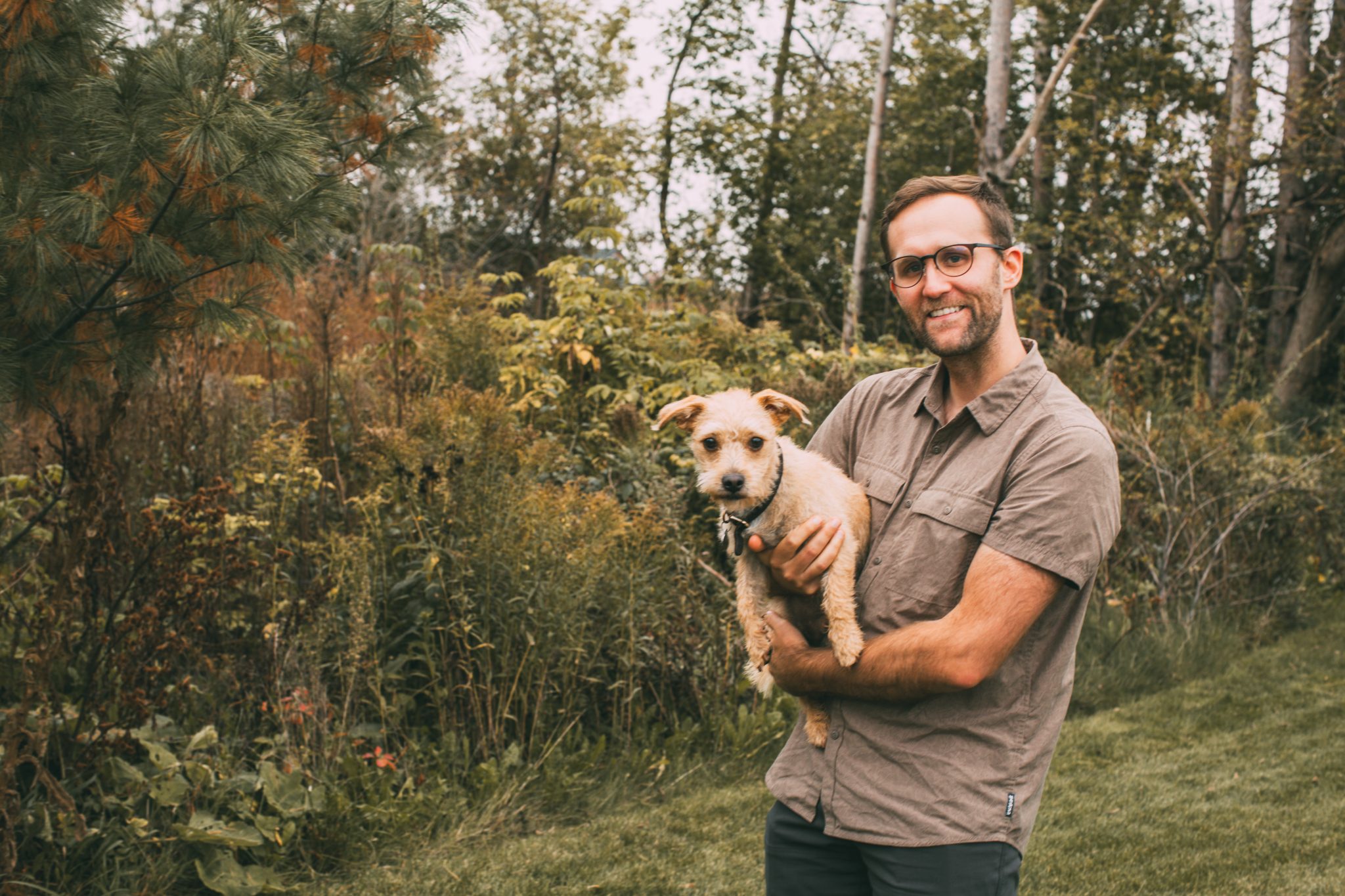 A man pictured holding a small dog in front of a grassy, wooded field. The man is wearing a light brown short-sleeved shirt and jeans.