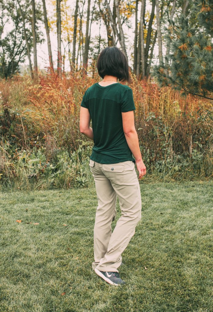 A woman poses in a grassy field, wearing tan hiking pants and a dark shirt, both from the sustainable travel clothing brand prAna.