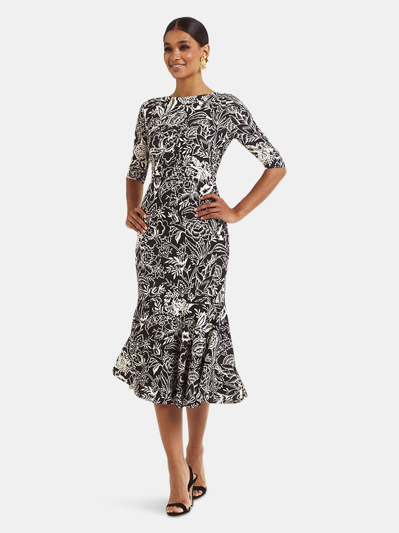 A darker skin woman models a Mermaid silhouette style dress with ruffle detail at hem. The dress has three-quarter length sleeves and has an all-over black and white floral pattern.