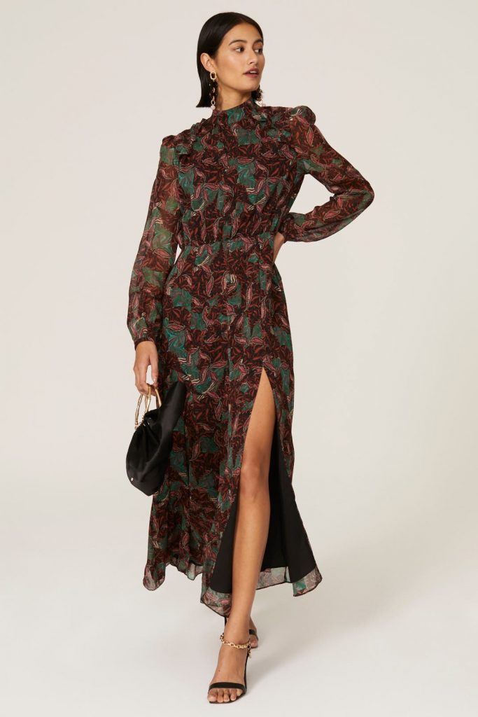 A young white female model wears a long brown dress with a peacock green pattern all over the dress. The dress has long sheet sleeves, a thigh-high slit, and mock-neck neckline.