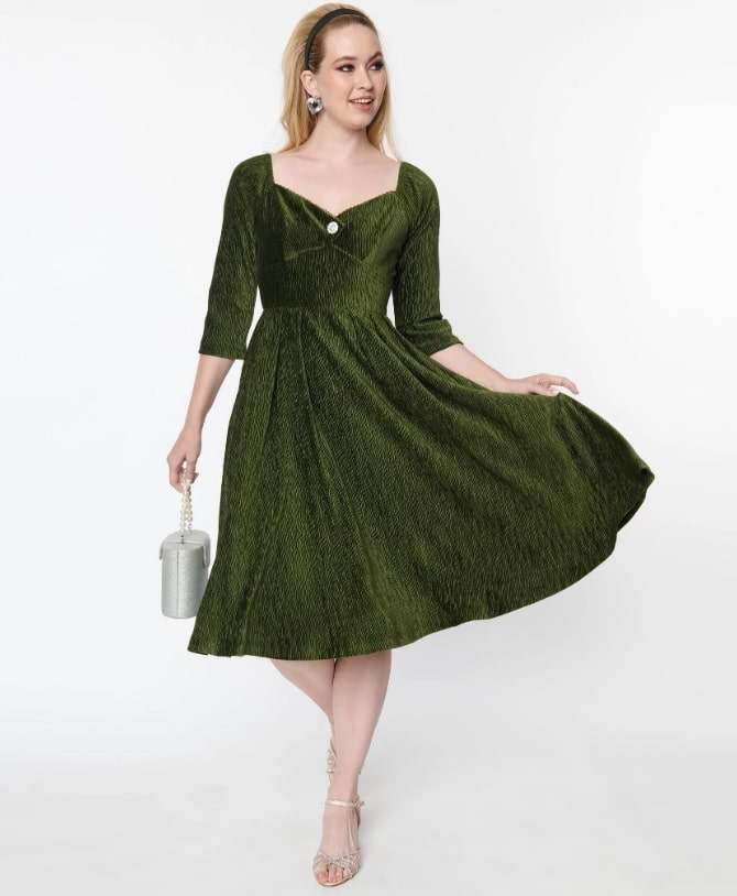 A white female model wears an olive green velvet dress. The dress is knee-length with three-quarter length sleeves and a sweetheart neckline.