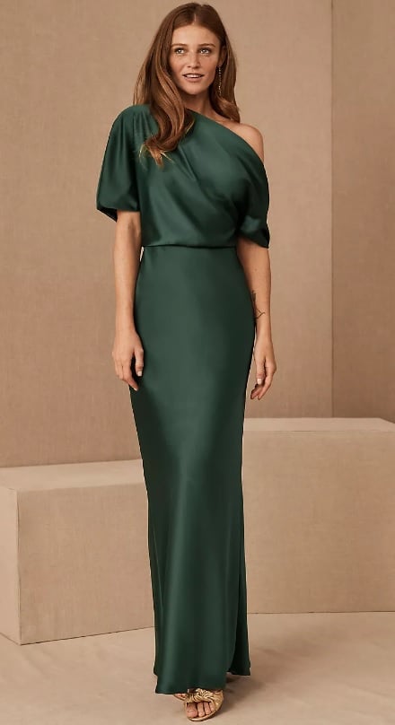 A young white model wears a one-shoulder emerald colored dress. The satin down has elbow-length sleeves and a neckline that drapes for an off-the-shoulder effect.