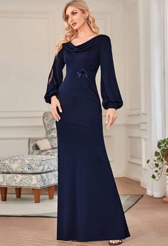 A white female model wears a floor length navy blue gown with sheer panel lantern sleeves. The dress has a sequin applique at the waist.