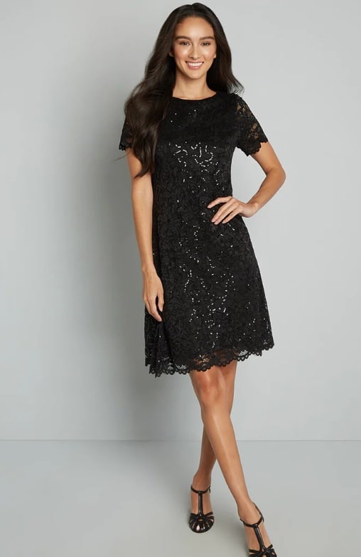 A white female model with long brown hair wears a knee-length black lace cocktail dress and black heels.