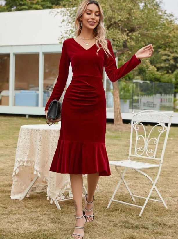 A white female model wears a knee-length deep red velvet dress. The dress has long sleeves, a v-neckline, and a ruffle hem.