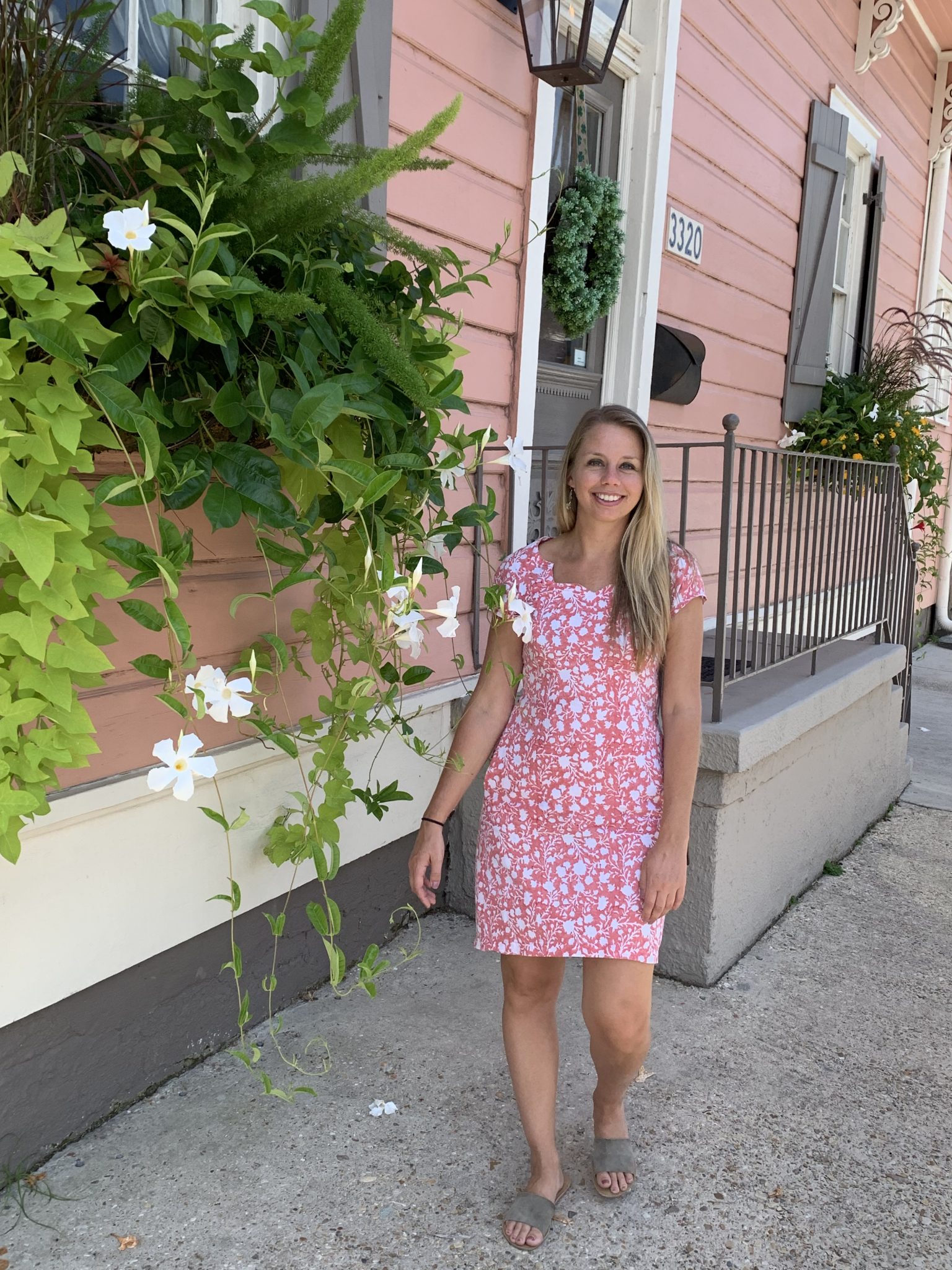 A woman poses on a sidewalk next to a light pink house with lush window planter boxes. She's wearing a summery pink floral dress.