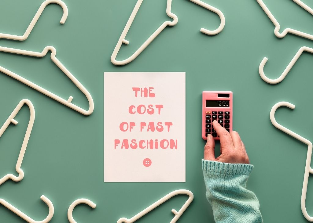 A flatlay image of plastic clothes hangers arranged on a green background. A person's hand rests on a small calculator, next to a piece of paper that reads "the cost of fast faschion" in bubbly pink text.