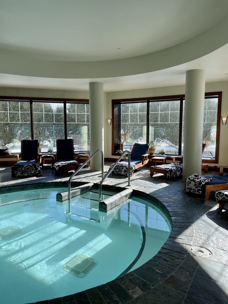 The spa area at the Aspira Spa in Elkhart Lake. The round pool is surrounded by comfortable padded pool chairs and towels. Windows line the walls of the room.