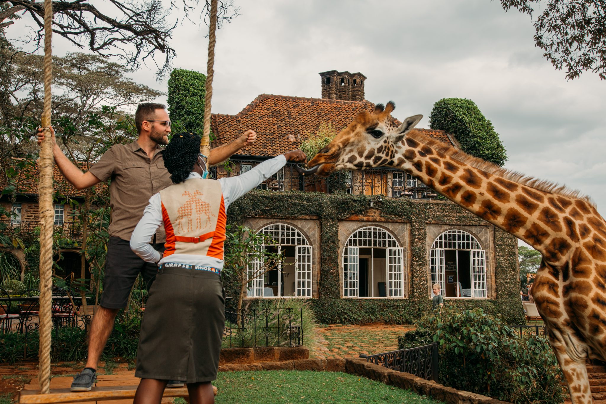 A man feeding a giraffe in front of Giraffe Manor and being guided by the staff.