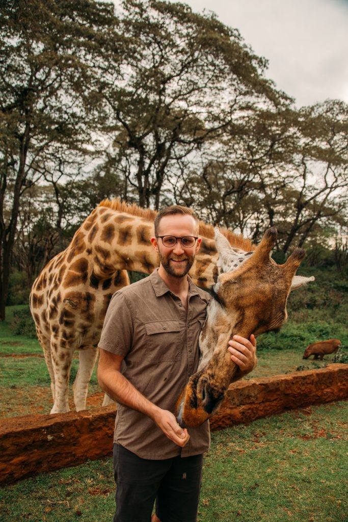 A man poses with a giraffe that's feeding from his hand. The man is wearing a brown short sleeved top and black shorts.