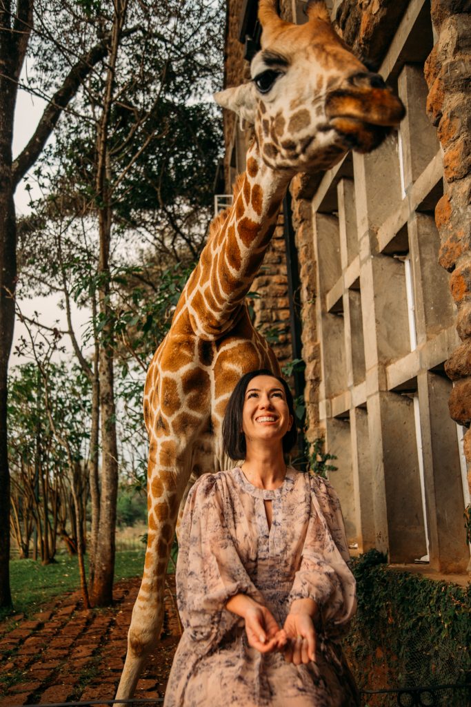 A woman is posing with a giraffe in front of a building.