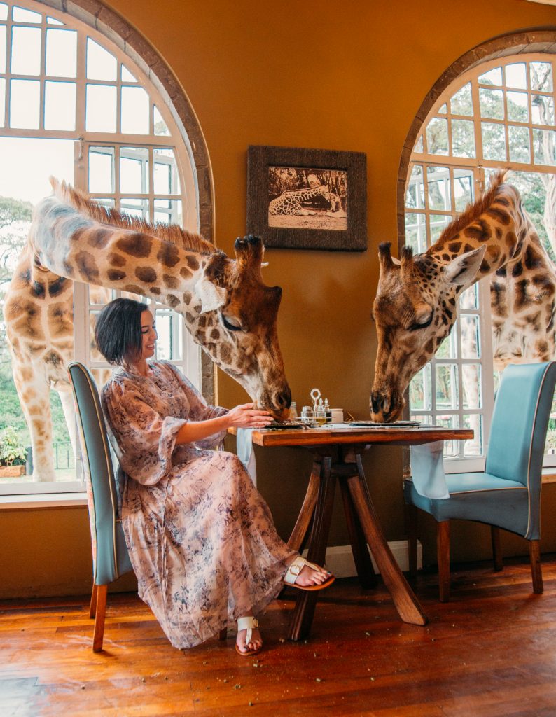 A woman in a light brown maxi dress sits at a small table for two, while two giraffes stick their heads through open windows, helping themselves to crumbs on the table