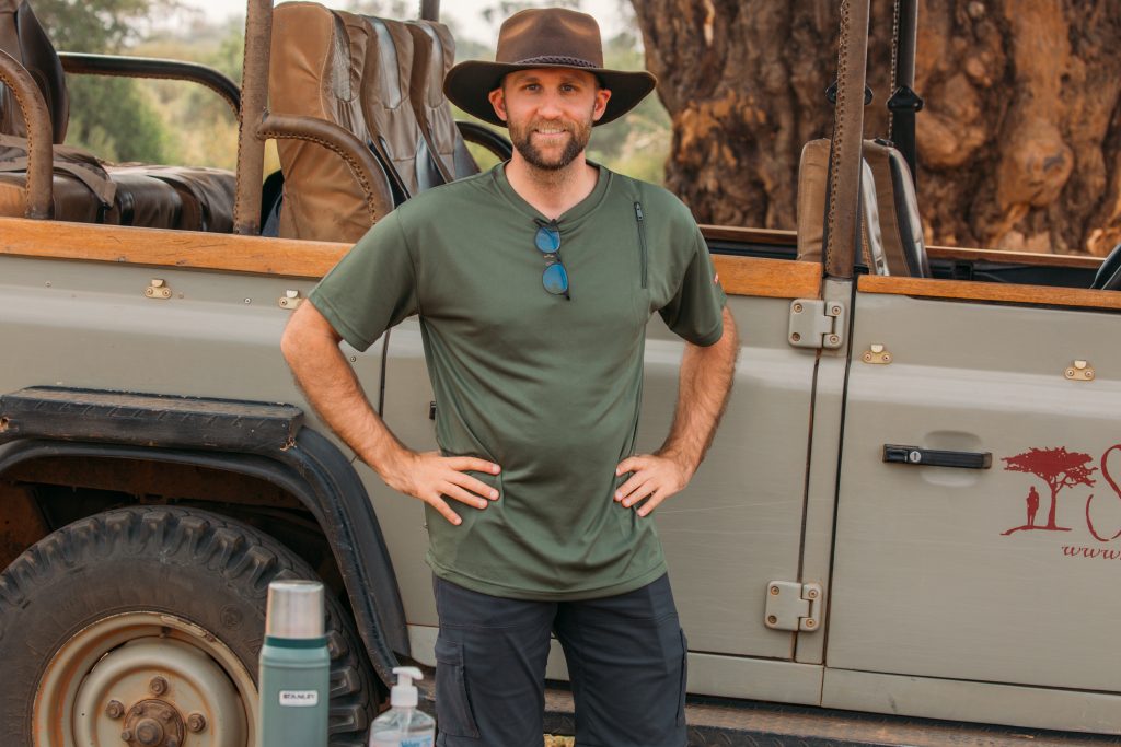 Zac wearing a SCOTTeVEST performance shirt in green and an oil skin hat, standing by a safari jeep in Kenya